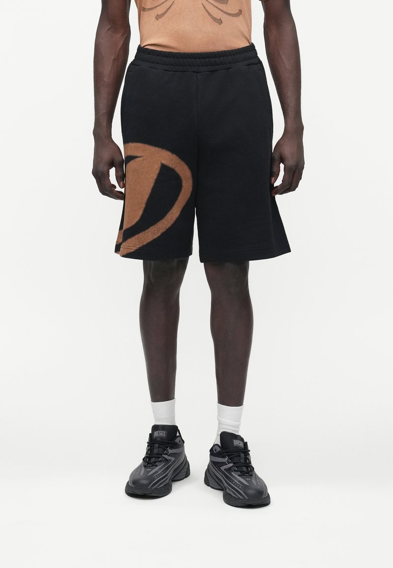 Persona de piel oscura que lleva pantalones cortos negros con un gran gráfico marrón en la pierna izquierda, calcetines blancos y zapatillas negras Diesel, de pie contra un fondo blanco.