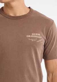 Bruine katoenen t-shirt met korte mouwen, geribbelde ronde hals en witte bedrukte tekst die "GUESS ORIGINALS 'Professional Rookie'" zegt.