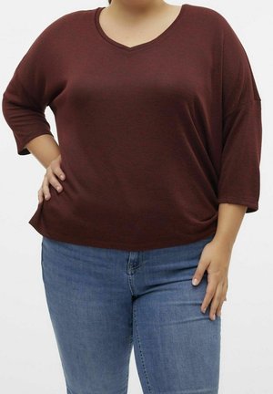 Vrouw draagt een bruin shirt met driekwart mouwen en blauwe spijkerbroek, met één hand op haar heup en de andere hand ontspannen langs haar zij.