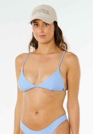 Kvinde iført lyseblå ribstrikket bikini og beige kasket med "RIP CURL"-logo, stående mod en ensfarvet hvid baggrund.