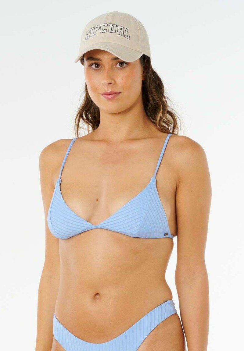 Donna che indossa un bikini a coste azzurro chiaro e un cappellino beige con logo "RIP CURL", in piedi davanti a uno sfondo bianco uniforme.