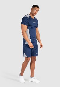 Marineblaues Sport-Shirt und Shorts-Set mit hellgrauen Seitenakzenten, figurbetontem Design und feuchtigkeitsableitendem Stoff. Weiße Turnschuhe ergänzen das Outfit.