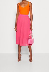 Top senza maniche arancione brillante con spalline regolabili, abbinato a una gonna rosa plissè. Il modello tiene in mano una piccola borsa rosa e indossa tacchi nudi.