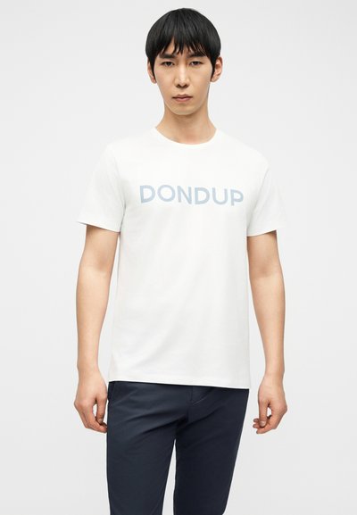 Άνδρας στέκεται μπροστά με λευκό μπλουζάκι με το κείμενο "DONDUP" και σκούρο μπλε παντελόνι σε απλό φωτεινό φόντο.