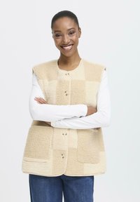 Bežs vilnas veste ar mīkstu un teksturētu apdari, kas aprīkota ar pogu aizdari un divām frontālām kabatām. Bez piedurknēm, apaļa kakla izgriezums.