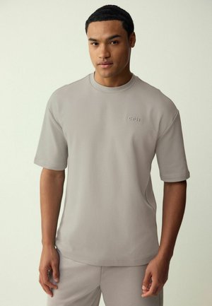 Young man wearing a light gray short-sleeve shirt and matching pants standing against a plain light background.