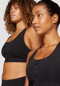 Os soutiens desportivos pretos com fecho frontal apresentam uma textura suave, alças largas e um design ajustado, usados por duas modelos com cabelo encaracolado e liso.