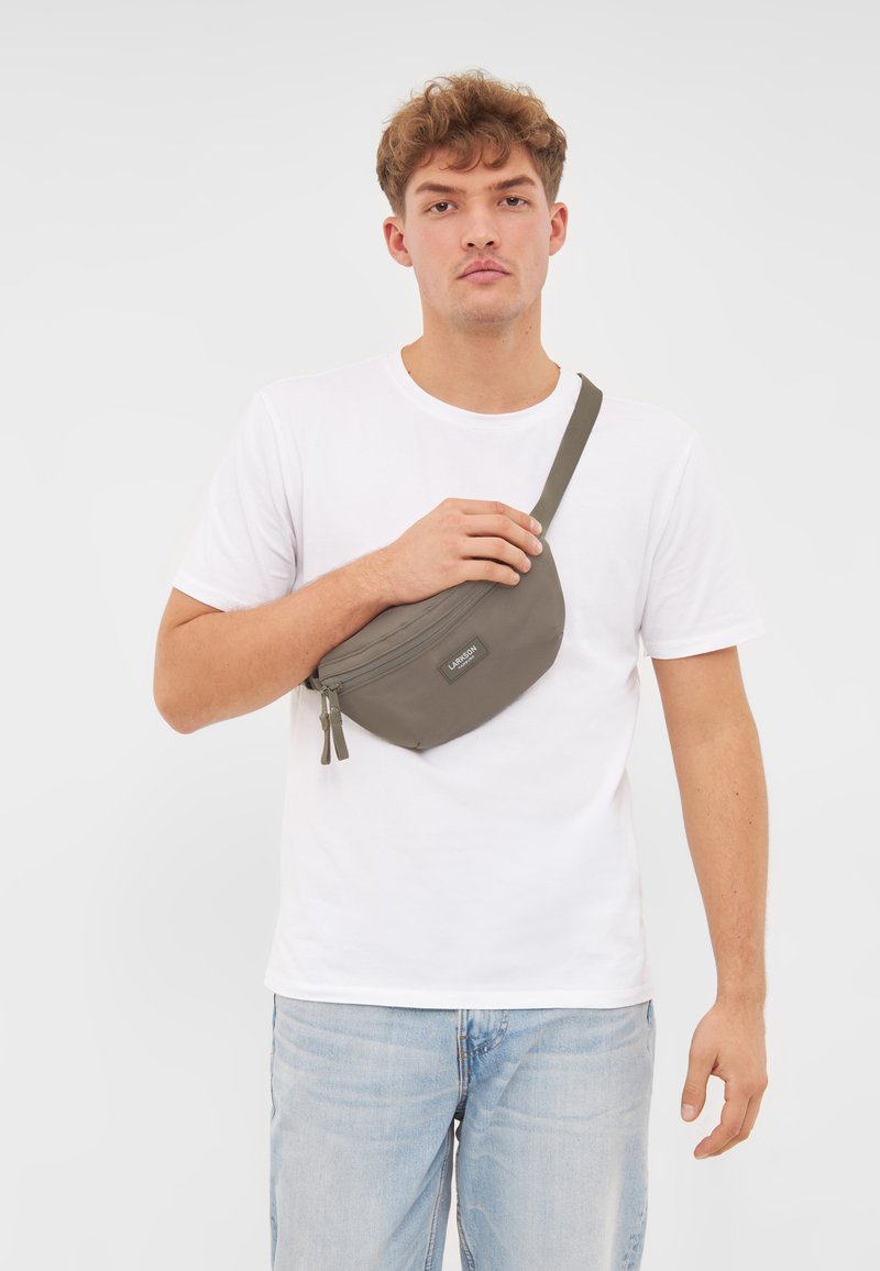 Olívazöld deréktáska puha anyagból, kerek formával, dupla cipzárral és állítható pánttal.