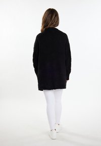 Lungo cardigan nero lavorato a maglia con una finitura texturizzata, apertura frontale e vestibilità morbida, abbinato a jeans skinny bianchi e sneakers bianche.