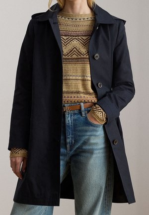Vrouw draagt een zwarte mantel met knopen over een beige gebreide trui met patroon, blauwe spijkerbroek en een bruine riem met een "RL" gesp, hand in zak.
