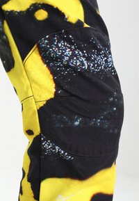 Svart och gult tyg med en texturerad mönstring. Har en sidoficka och synlig sömnad. Designen innehåller abstrakta former och färger.