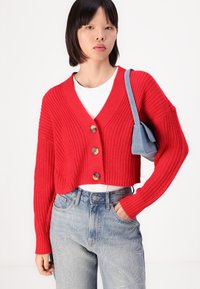 Rode cropped cardigan met een gestructureerde brei, met drie grote knopen en een V-hals ontwerp, gecombineerd met lichtblauwe jeans.