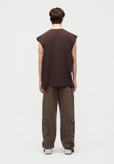 Άνδρας γυρισμένος από την πλάτη, φορά άσπρο σκούρο καφέ αμάνικο πουκάμισο και χαλαρό καφέ cargo παντελόνι με φαρδιά πόδια και λευκά παπούτσια με τετράγωνη μύτη.