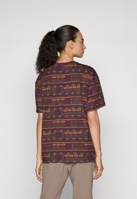 Short-sleeve shirt in dark maroon fabric featuring geometric patterns in yellow, green, and blue. Crew neckline and relaxed fit.