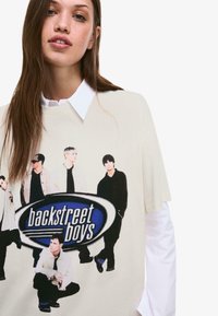 Kremowy, oversize'owy t-shirt z graficznym nadrukiem logo Backstreet Boys oraz wizerunkami członków zespołu w różnych pozach.