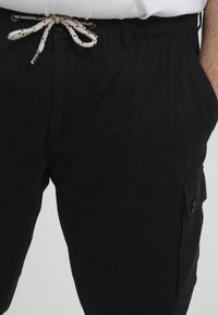 Schwarze Cargo-Hosen mit elastischem Bund und Kordelzug, ausgestattet mit Seitentaschen und einer mit Knopf verschließbaren Cargo-Tasche am rechten Oberschenkel.