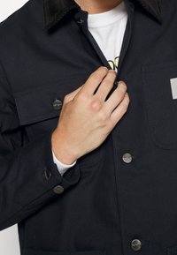 Dunkelblaue Jacke mit einem Cordkragen, ausgestattet mit Druckknopfverschlüssen und Fronttaschen. Der Stoff wirkt strukturiert und strapazierfähig.