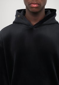 Felpa nera con una texture morbida e liscia, dotata di cappuccio regolabile con cordino e un piccolo logo dorato vicino all'collo.