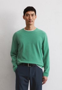 Grüner Strickpullover mit strukturiertem Muster und weißen Rundhalsausschnitt, kombiniert mit navyblauen Hosen und einem schwarzen Gürtel. Einfaches Design, lange Ärmel.
