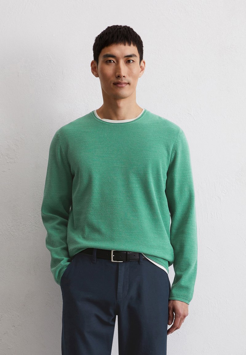 Grüner Strickpullover mit strukturiertem Muster und weißen Rundhalsausschnitt, kombiniert mit navyblauen Hosen und einem schwarzen Gürtel. Einfaches Design, lange Ärmel.