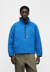 Μπλε αδιάβροχο μπουφάν με ψηλό γιακά, μπροστινό φερμουάρ μέχρι τη μέση και λογότυπο κεντημένο. Ελαφρύ ύφασμα με άνετη εφαρμογή και ελαστικές μανσέτες.