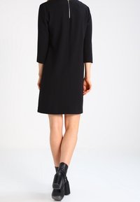 Musta polvipituinen mekko, jossa on kolme neljäsosaa hihat, takana vetoketjukiinnitys. Yhdistetty muovikorkoisiin mustiin saappaisiin.