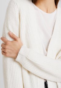 Biały sweter z dzianiny z dekoltem w szpic, prążkowaną fakturą i długimi rękawami. Ręka modelki opiera się na rękawie, podkreślając jego dopasowanie.