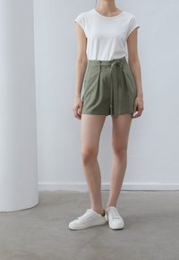 Weißes, enges T-Shirt kombiniert mit olivgrünen, hochtaillierten Shorts mit Bindegürtel. Die Person trägt weiße Sneaker und steht auf einem Betonboden.