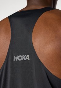 Débardeur de sport noir avec un design dos nageur, fabriqué dans un matériau respirant, avec un logo "HOKA" gris réfléchissant au dos.
