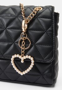 Sac à main en cuir noir matelassé avec une bandoulière en chaîne dorée, ornée d'un pendentif en forme de cœur avec des perles et de détails en métal doré.