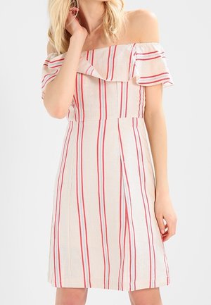 Mujer con vestido de rayas verticales rojas y blancas fuera del hombro, de pie con una mano cerca del rostro y la otra relajada a un lado.