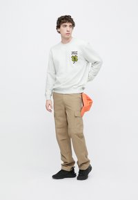 Hellgrauer Pullover mit grünem Kleeblatt-Design, kombiniert mit beigen Cargo-Hosen mit Seitentaschen. Das Modell trägt schwarze Turnschuhe und eine orangefarbene Kappe.