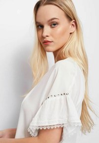 Blusa bianca con rifiniture in pizzo sulle maniche e borchie metalliche sulle spalle. Il tessuto è liscio, con una vestibilità comoda e scollatura ampia.