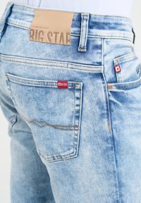 Šviesiai mėlynos džinsinės kelnės su tan spalvos odiniu įsiuvu liemens srityje ir raudonais logotipo ženklais. Matoma siūlų ir kišenių detalizacija.