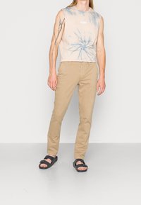 Ljus beige ärmlös skjorta med blått tie-dye mönster, tanbyxor och svarta sandaler med remdesign. Modellen står vänd framåt.