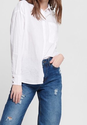 Personne portant une chemise blanche à manches longues boutonnée rentrée dans un jean délavé bleu taille haute, la main dans la poche.