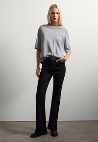Donna con lunghi capelli biondi che indossa una maglietta grigia larga infilata in pantaloni neri a vita alta, in piedi con una mano nella tasca.