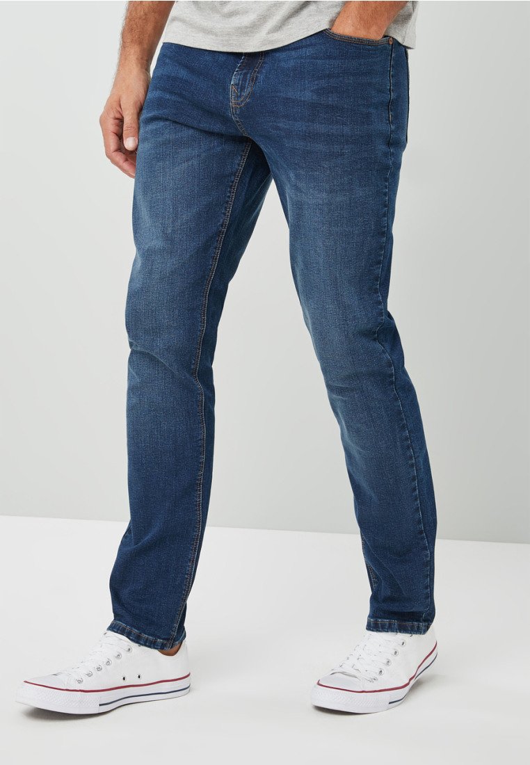 Jeans de denim azul oscuro con un corte slim, que cuentan con costuras cosidas y un diseño clásico de cinco bolsillos, combinados con zapatillas blancas.