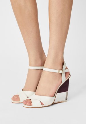 Sandales compensées en cuir blanc avec bride à la cheville, talon en bois clair et foncé, design à bout ouvert, et sangles supérieures croisées.