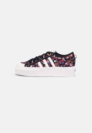 Lavskoet sneaker med sort stof øvre, flerfarvet blomsterprint, hvide striber, tyk hvid gummisål og sorte snørebånd på ensfarvet baggrund.