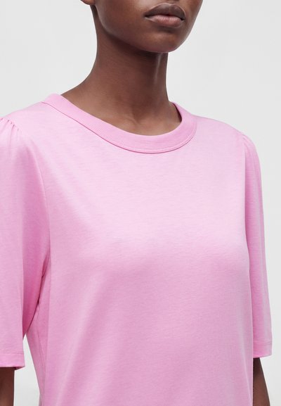 Gros plan d'une personne portant un t-shirt rose clair à manches courtes avec un col rond, sur un fond clair uni.