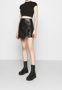 T-shirt noir court associé à une jupe portefeuille en simili cuir noir avec une poche zippée sur le côté et des bottines à plateforme lacées noires.