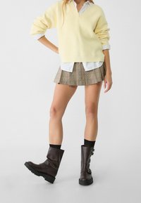 Żółty sweter w prążki, biały koszula z kołnierzykiem, mini spódniczka w kratę oraz brązowe skórzane botki na kostkę z detalami w postaci klamerek.