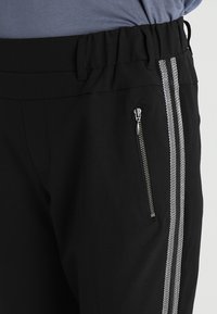 Zwarte sportbroek met een elastische tailleband, voorzien van een ritszak en twee verticale witte strepen langs de zijkant. Zachte stoftextuur.