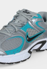 Γκρι και τιρκουάζ αθλητικό παπούτσι Nike με διχτυωτά πάνελ, μαύρο λογότυπο Swoosh και λευκή σόλα, γωνιακή λήψη που δείχνει την εξωτερική πλευρά και τα κορδόνια.