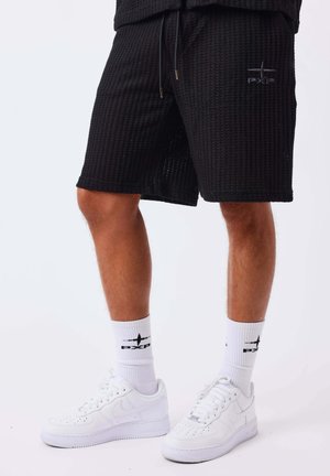 Uomo che indossa pantaloncini neri strutturati, calzini bianchi con logo e scarpe da ginnastica bianche su uno sfondo chiaro uniforme.