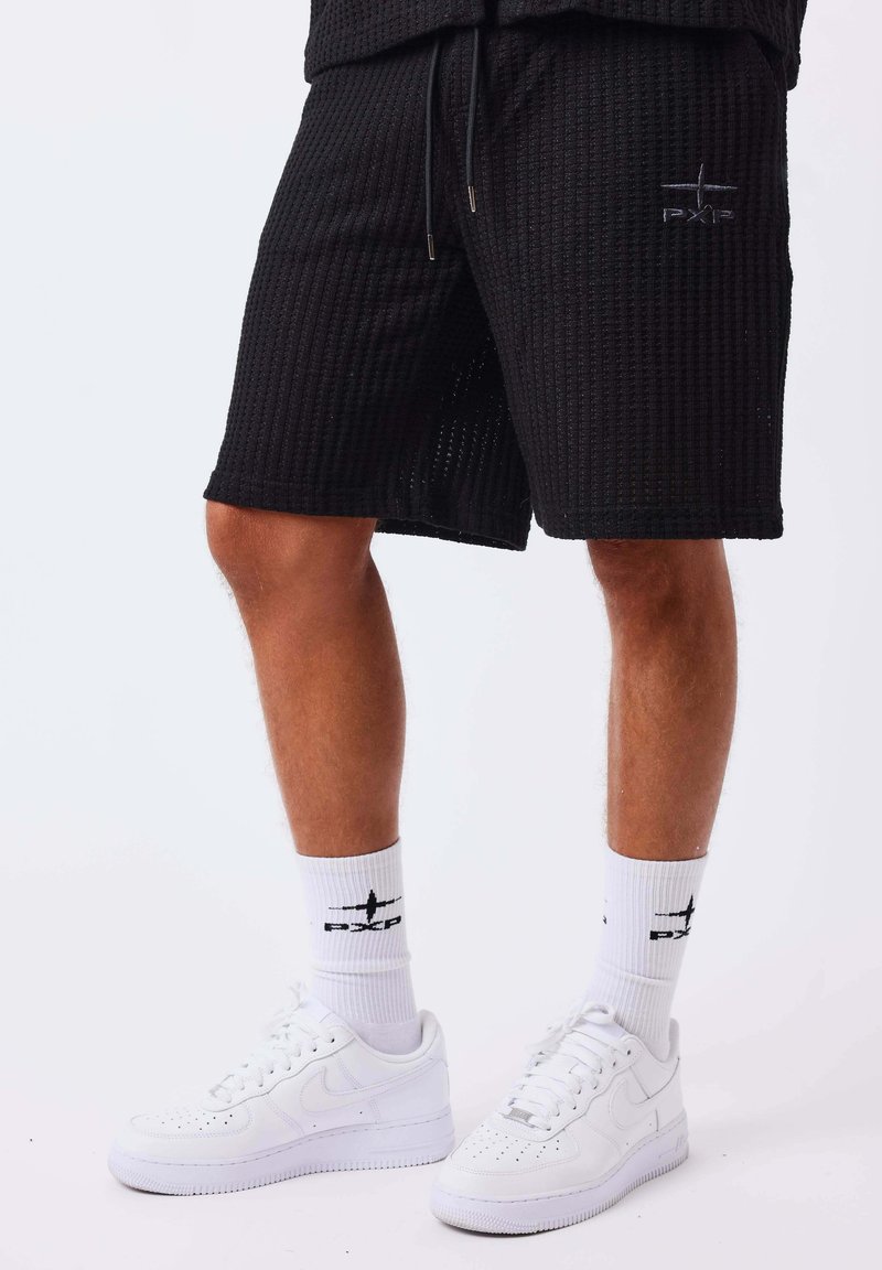 Homme portant un short noir texturé, des chaussettes blanches avec logo, et des baskets blanches sur un fond clair uni.