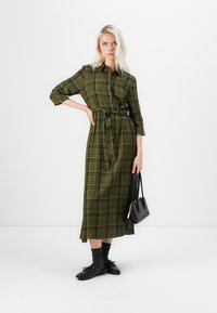 Green plaid button-up dress with a belted waist, three-quarter sleeves, and a mid-length hem. Black bag and shoes complete the outfit.