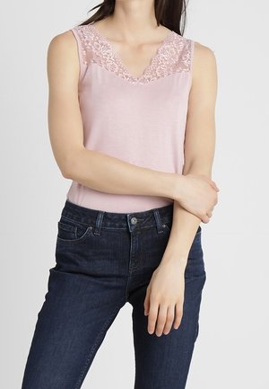 Femme portant un débardeur rose clair avec une bordure en dentelle et un jean bleu foncé, debout les bras croisés à la hauteur de la taille.