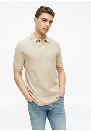 Junger Mann mit kurzem lockigem Haar, trägt ein beiges kurzärmliges Polohemd und hellblaue Jeans, Hände in den Taschen, neutraler Gesichtsausdruck, weißer Hintergrund.